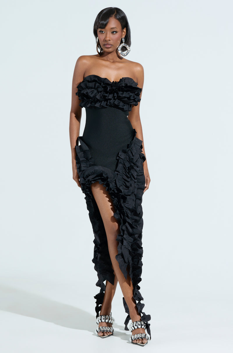 A woman models the CITY LIGHTS RUFFLE MIDI DRESS, a strapless gown with ruffle trim and an asymmetrical hemline. She accessorizes with large statement earrings and metallic open-toed heels against a plain white background.