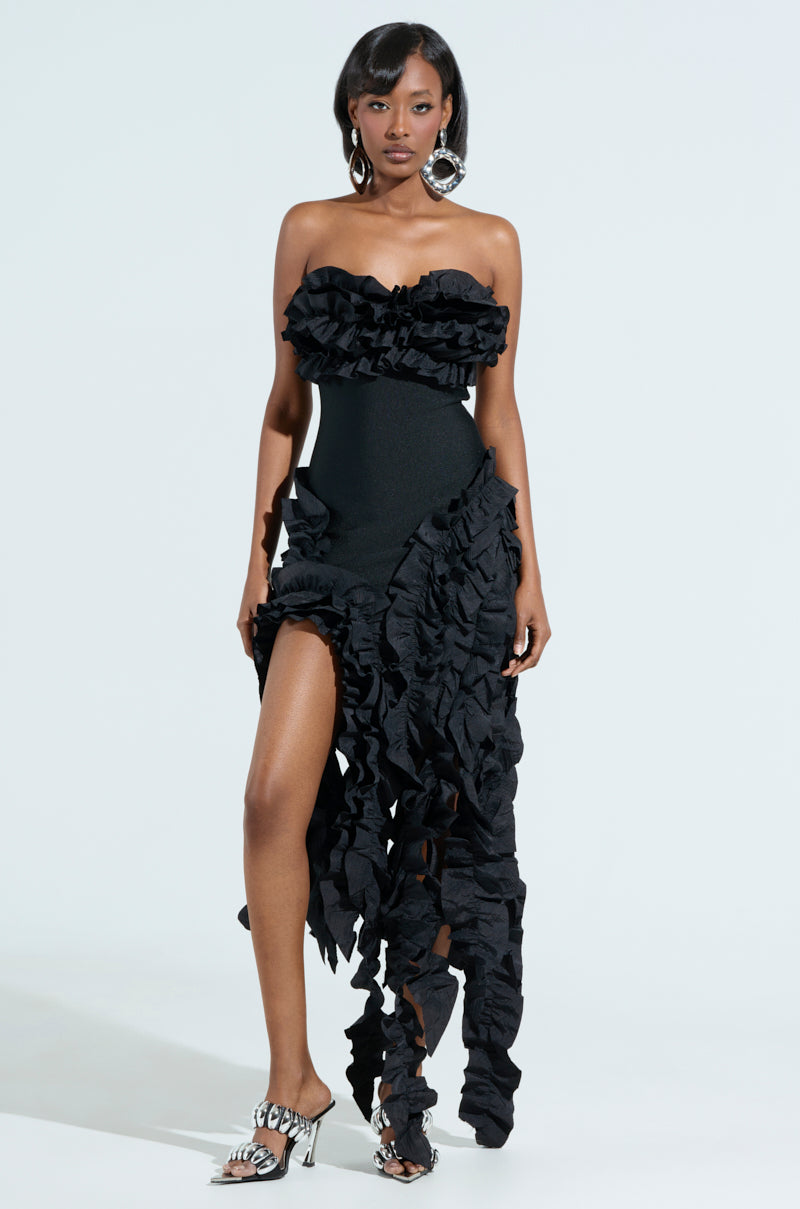 A woman models the CITY LIGHTS RUFFLE MIDI DRESS, a strapless gown with layered ruffles, an asymmetrical hem, and a high slit. Elegantly paired with large silver earrings and black-and-white patterned heels, she stands against a plain white background.