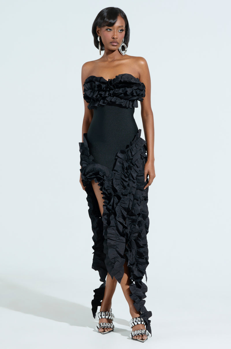A woman poses on a white background in the CITY LIGHTS RUFFLE MIDI DRESS, showcasing its strapless design with a bandage bodice, elaborate ruffles, and high slit. She has short, straight black hair, large circular earrings, and ornate high-heeled shoes.