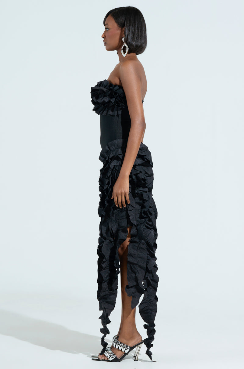 A woman in the CITY LIGHTS RUFFLE MIDI DRESS, featuring a black ruffled strapless design with a bandage bodice, stands against a white background. The dress has an elongated back, paired with silver heels. Her hair is straight and shoulder-length, complemented by large hoop earrings.