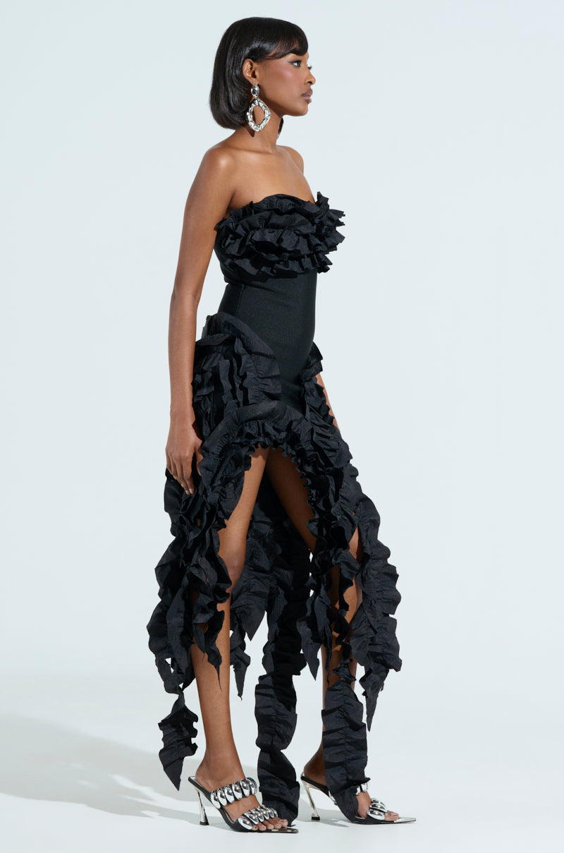 The person is wearing the CITY LIGHTS RUFFLE MIDI DRESS, a strapless gown with ruffled floor-length layers and a bandage bodice enhancing their elegant silhouette. They accessorize with large statement earrings and open-toed silver shoes against a plain white background.