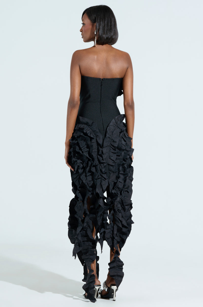 A woman with short black hair poses with her back turned, wearing the CITY LIGHTS RUFFLE MIDI DRESS featuring a strapless bandage bodice and cascading ruffles. She completes her look with silver earrings and black high heels, set against a plain white background.
