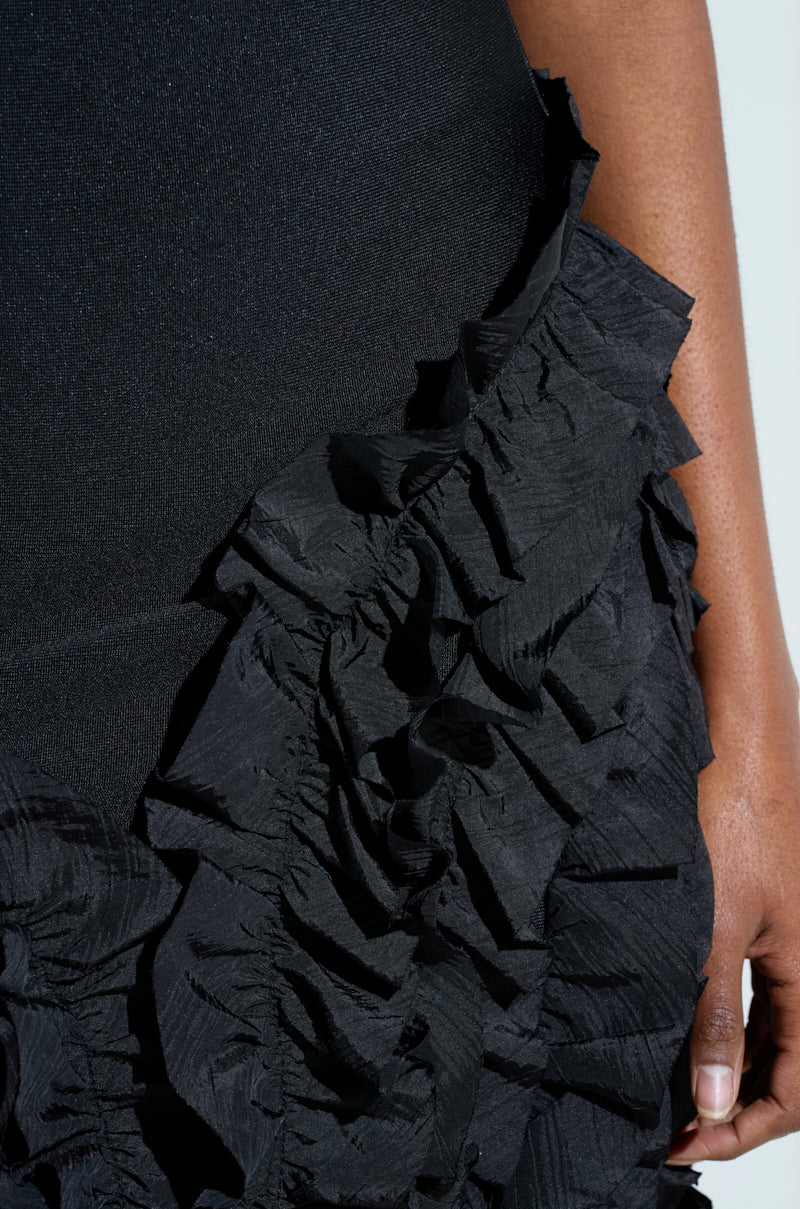 Close-up of a person in the CITY LIGHTS RUFFLE MIDI DRESS, a textured black garment featuring ruffle trim. The crinkled fabric adds depth, and the bandage bodice enhances their profile. The person's hand is partially visible on the right edge.