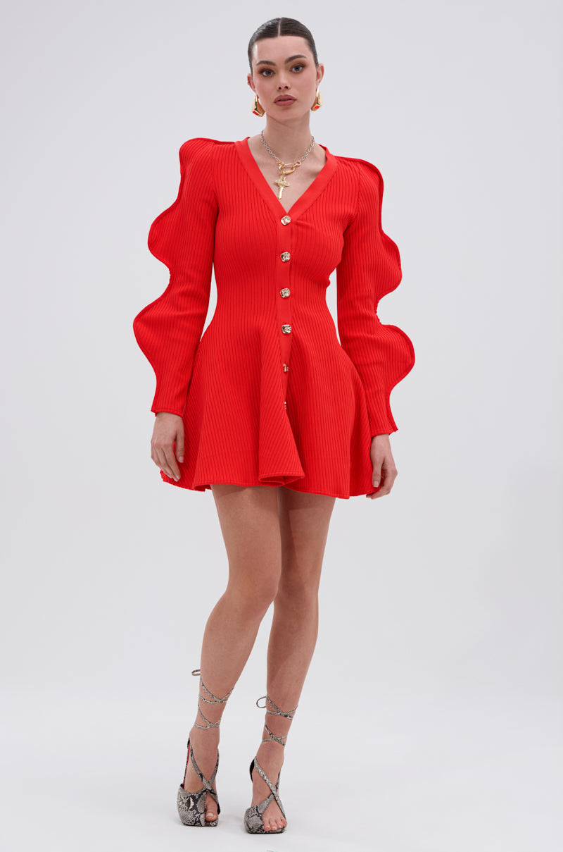 A woman models the COOLEST ON THE BLOCK MINI DRESS IN RED—a bright red ribbed knit mini dress with dramatic ripple sleeves—styled with gold jewelry and strappy snakeskin heels, set against a plain light gray background.