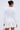 A woman with her hair in a bun is photographed from behind, wearing the COOLEST ON THE BLOCK MINI DRESS—a white mini with long sleeves and wavy side details. She accessorizes with a small bag and large pearl earrings against a light background.