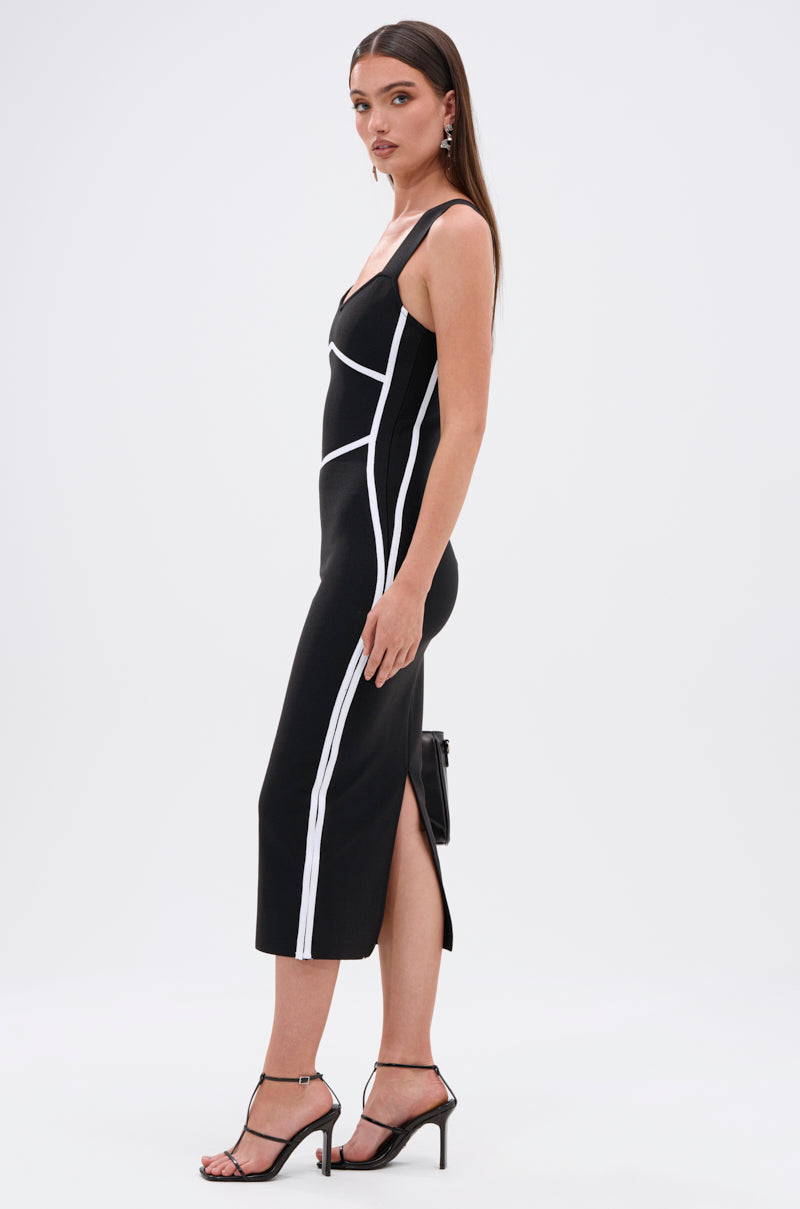 A woman stands in profile against a white background wearing the ALL ABOUT THAT BODY BANDAGE MIDI DRESS—a fitted, sleeveless black midi with white geometric paneling and a side slit—styled with hoop earrings, straight hair, and black high-heeled sandals.