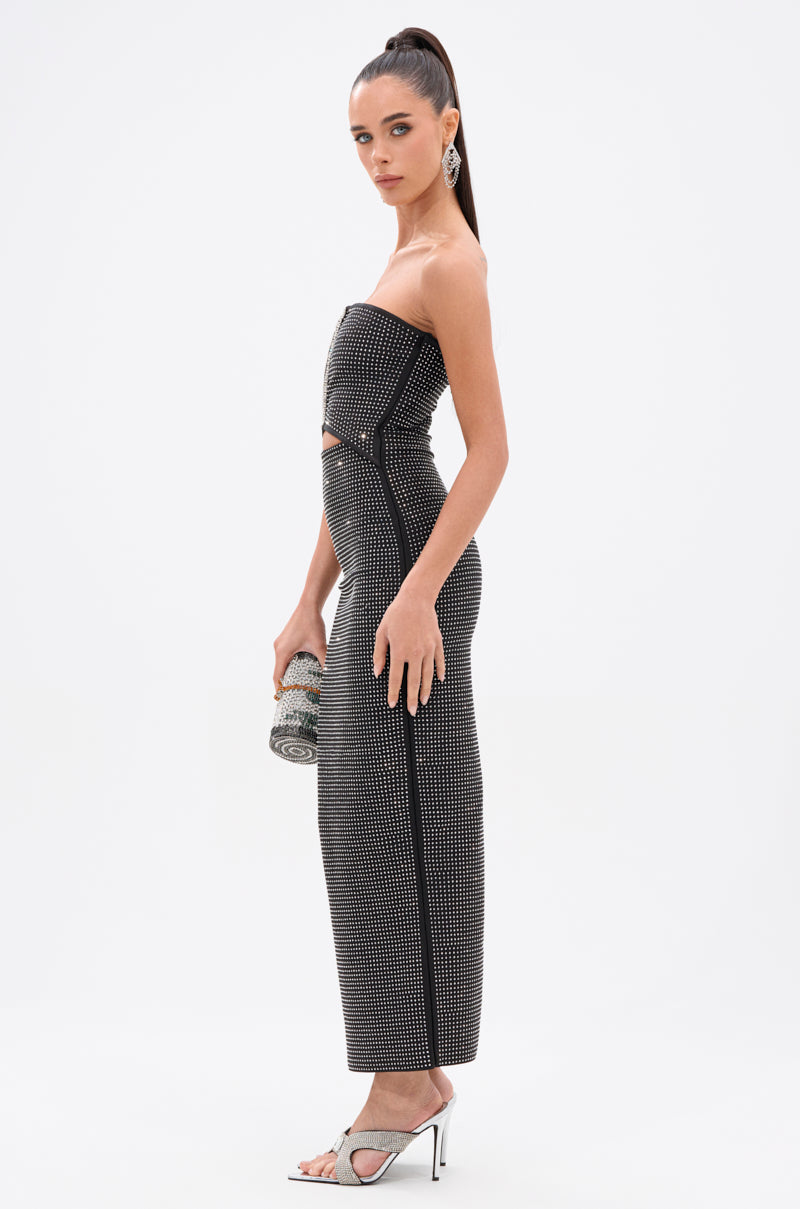 A woman stands sideways against a white background, wearing the ALINA RHINESTONE MIDI DRESS with small studs, high heels, statement earrings, and holding a rhinestone clutch. Her hair is styled in a high ponytail.