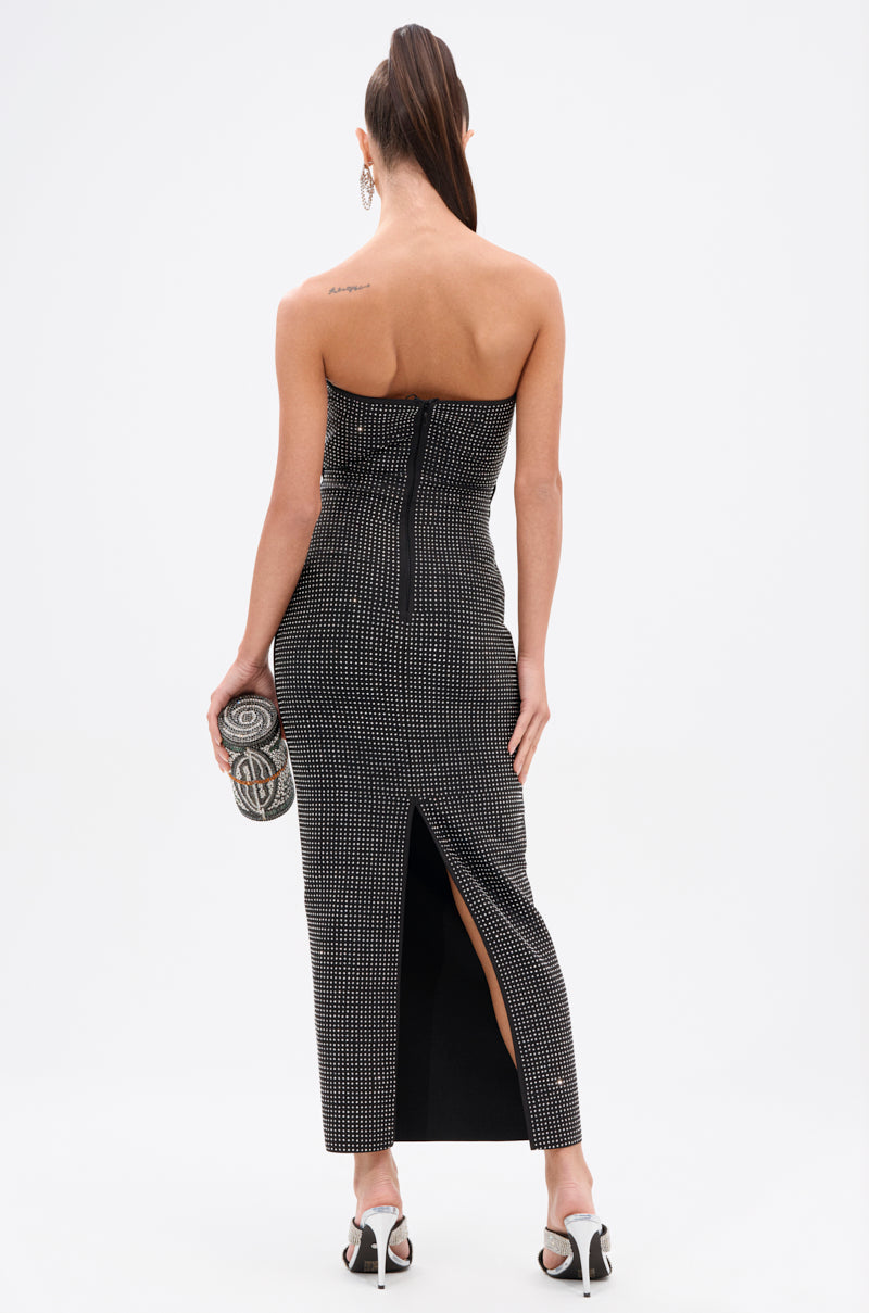 A woman stands facing away in the ALINA RHINESTONE MIDI DRESS, a fitted black strapless bodycon with silver rhinestone accents and a back slit. She pairs it with white high-heeled sandals, a patterned clutch, and wears her hair in a high ponytail.