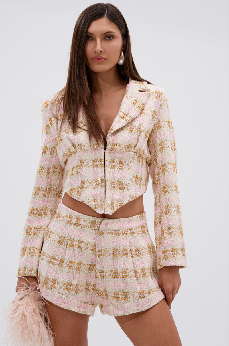 A woman poses against a plain background in the FLORENCE TWEED BLAZER—a pastel pink, cropped jacket with subtle plaid and feminine details—paired with matching high-waisted shorts and holding a small pink feathered accessory. 0