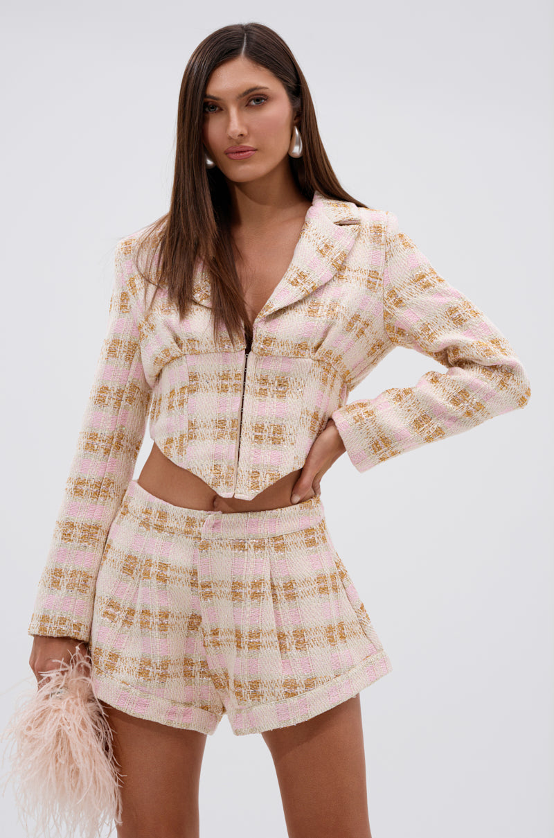 A woman stands confidently in the FLORENCE TWEED BLAZER with matching high-waisted shorts, holding a pale feathered clutch. She wears large hoop earrings and has long, straight hair against a plain light background. 1