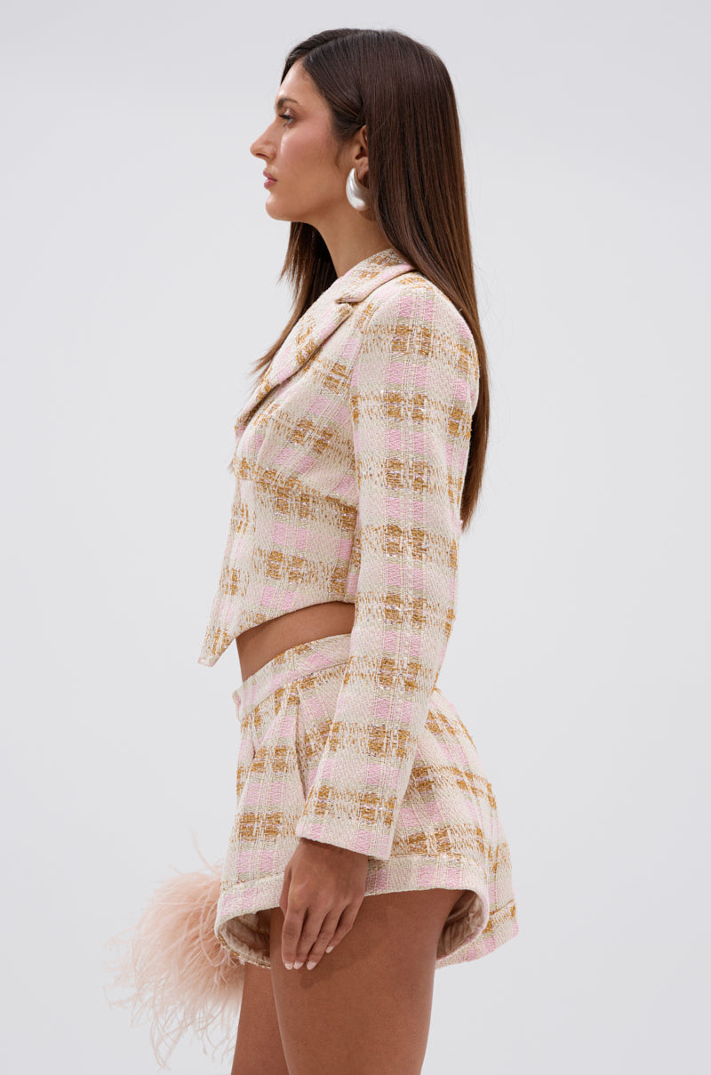A woman with long brown hair in profile wears the FLORENCE TWEED BLAZER in pastel pink, paired with matching high-waisted shorts, large round earrings, and holding a pink feather accessory against a plain light background. 2