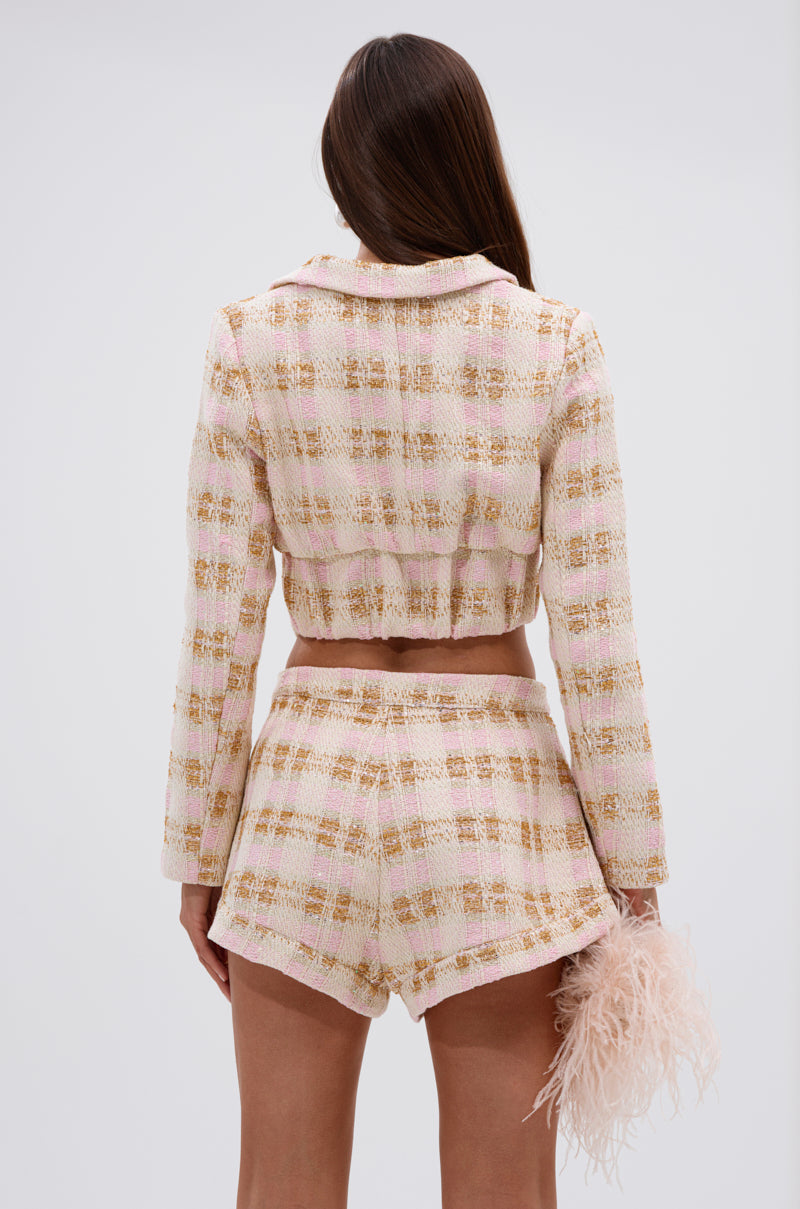 A woman faces away from the camera wearing the FLORENCE TWEED BLAZER in pastel pink with matching high-waisted shorts. She holds a pale pink feathered clutch in her right hand against a plain white background. 3