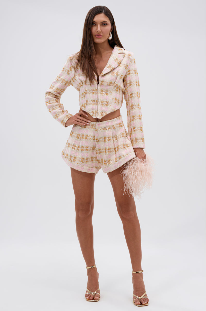 A woman wears the FLORENCE TWEED BLAZER in pastel pink, paired with gold high-waisted shorts, gold high-heeled sandals, and a pale pink feathered clutch against a plain white background. 5