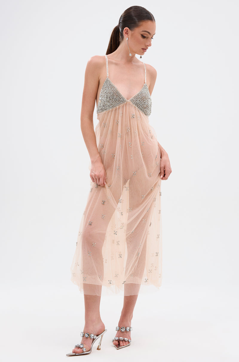 Wearing the DO YOU BELIEVE IN LOVE RHINESTONE MESH MIDI DRESS, a woman stands gracefully in its sheer nude fabric and sparkling embellishments, paired with metallic heels and her hair tied back as she gazes down, one hand resting on the dress.
