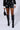 A person is sporting the IN THE DARK FAUX LEATHER SHORTS in high-waisted black faux leather, paired with a black top featuring sheer sleeves and complemented by sleek black knee-high boots. They are carrying a chic black handbag at their side against a plain, light grayish-white backdrop.