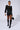 A person poses confidently against a plain background, wearing a black sheer long-sleeve blouse with white cuffs and collar, the IN THE DARK FAUX LEATHER SHORT, black knee-high boots, and large hoop earrings. They hold a small black handbag and have a serious expression.