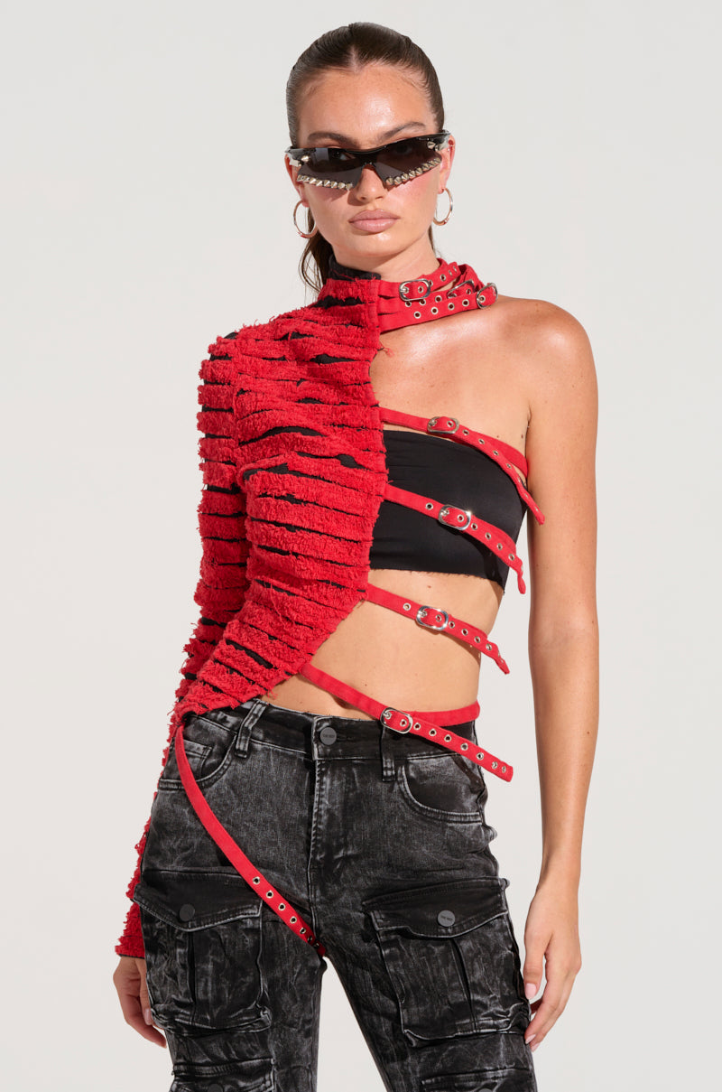 A person wearing the PRETTY RECKLESS DISTRESSED DENIM HALF THERE TOP, known for its distinctive red texture and single sleeve design adorned with multiple buckle straps, paired with stylish black cargo pants. They have their long hair tied back, accessorized with chic black sunglasses and hoop earrings, while posing against a plain background in a striking color block style.