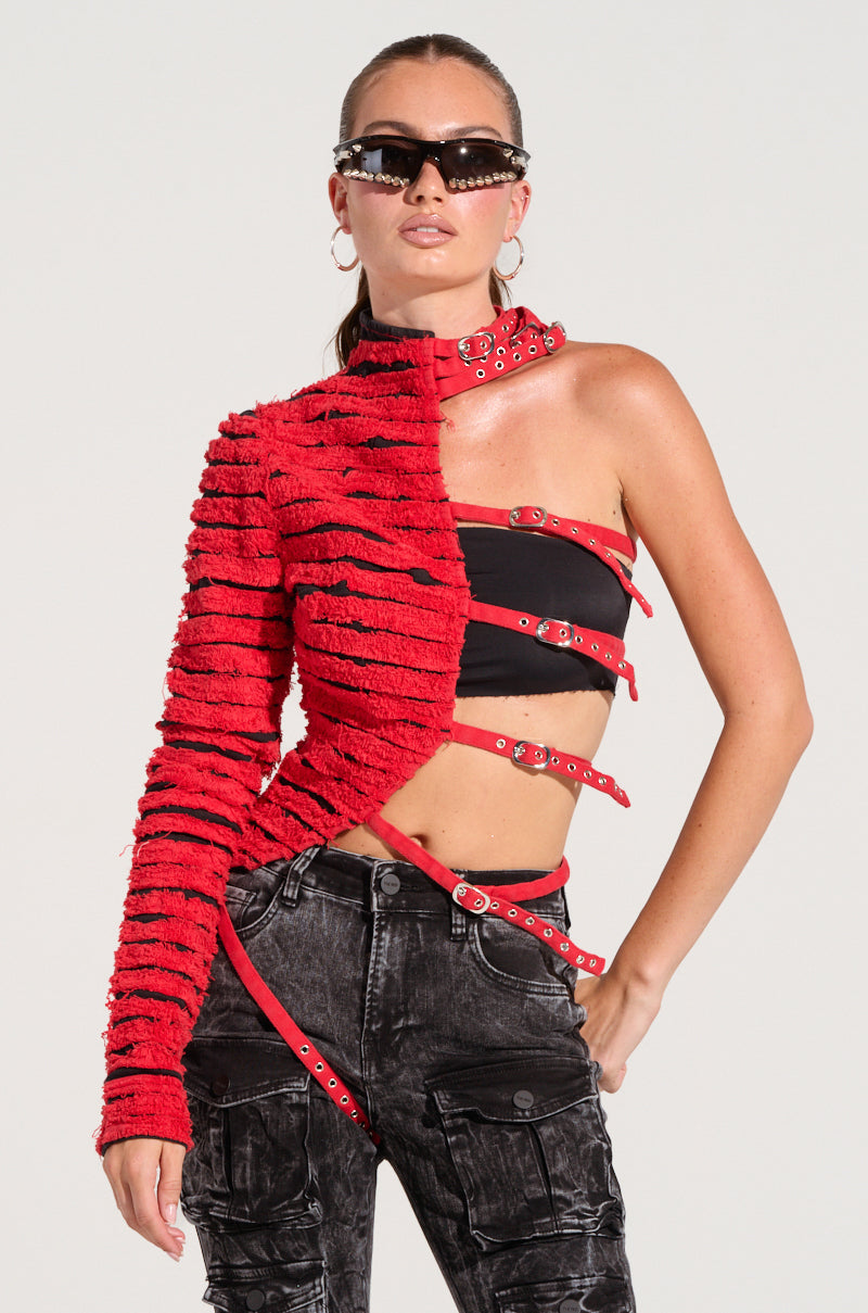 A person stands confidently against a plain background, wearing the PRETTY RECKLESS DISTRESSED DENIM HALF THERE TOP in red—a textured, one-sleeve top with multiple straps on the other side—paired with black distressed denim pants with pockets and stylish sunglasses.