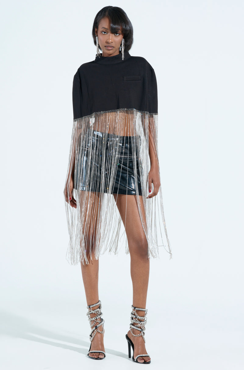 A person confidently wears a NEW LEVEL SHINE CROPPED BLOUSE with long silver rhinestone fringe and black leather shorts, complemented by strappy silver high heels and large hoop earrings, against a plain white background.