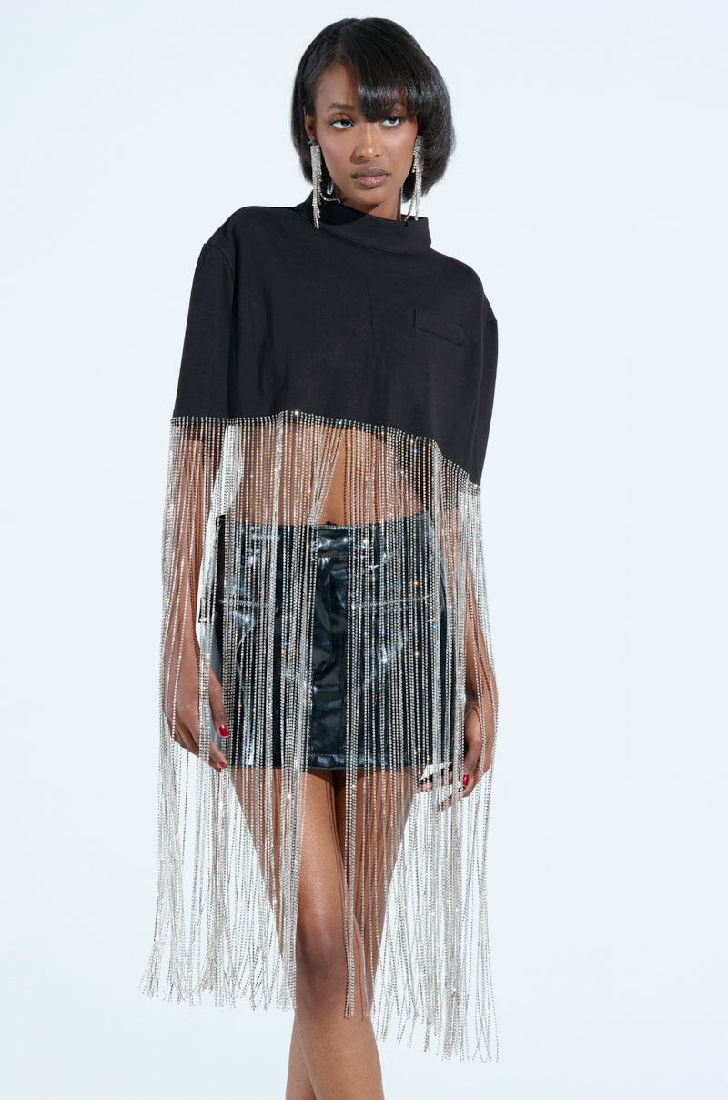 A person confidently poses in a stylish outfit with the NEW LEVEL SHINE CROPPED BLOUSE featuring rhinestone fringe and shiny black shorts, shoulder-length hair, statement earrings, and a chic cropped cape against a plain light background.