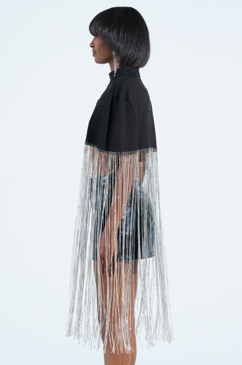 A person with short dark hair is wearing the NEW LEVEL SHINE CROPPED BLOUSE, adorned with long silver rhinestone fringe cascading to knee-length. They exude an ultra-glam vibe while facing left against a plain white background.