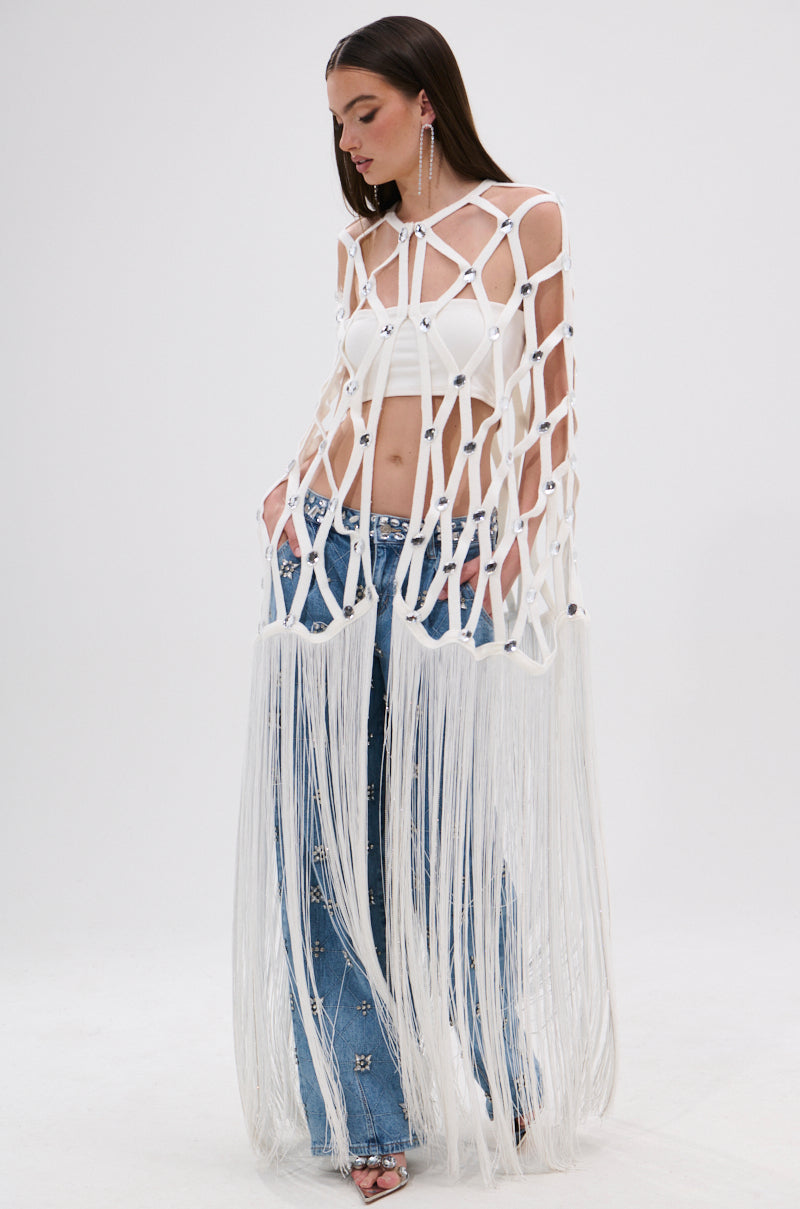 A woman wears a white cropped top, blue embellished jeans, and the dramatic WHITE ZELINA FRINGE EMBELLISHED LONG CARDIGAN, standing against a plain white background.