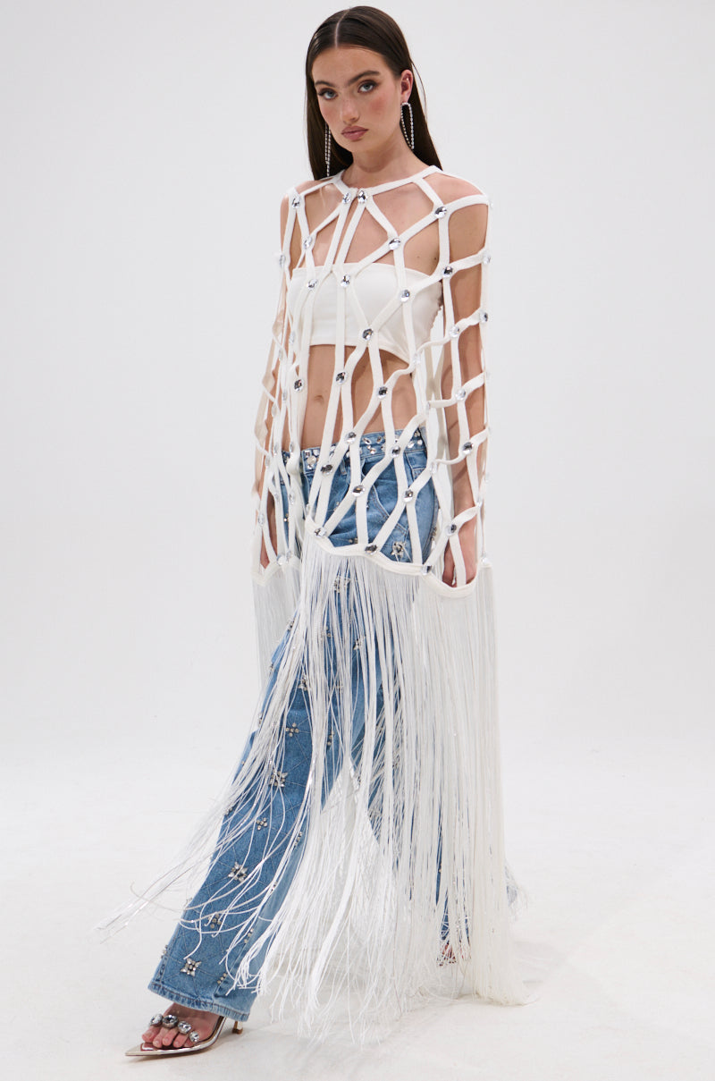 A woman with straight brown hair, wearing hoop earrings, a white crop top, mirrored gem embellished blue jeans, open-toed heels, and a WHITE ZELINA FRINGE EMBELLISHED LONG CARDIGAN walks forward against a plain white background.