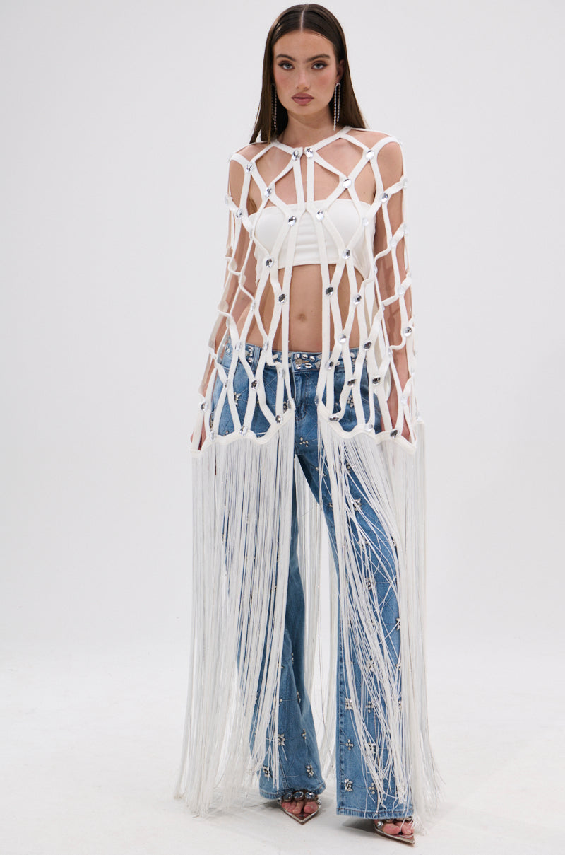 A woman stands facing forward in a white crop top, blue flared jeans with mirrored gems, and the WHITE ZELINA FRINGE EMBELLISHED LONG CARDIGAN. Her straight brown hair and neutral look create a perfect festival outfit against a plain white background.