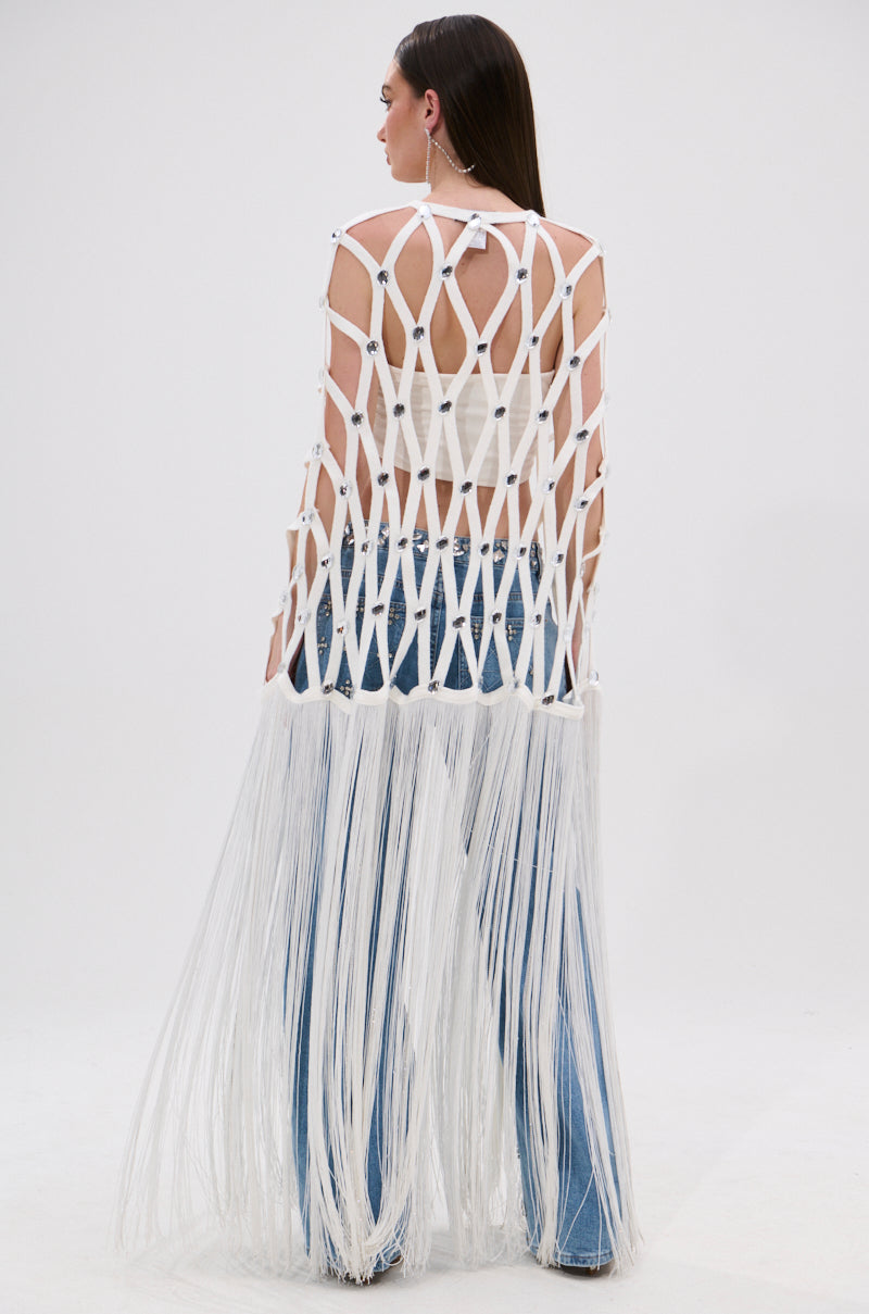 A woman faces away, wearing the WHITE ZELINA FRINGE EMBELLISHED LONG CARDIGAN—a festival-ready netted shawl with diamond patterns and long fringe draping below her knees, styled over a white top and blue jeans.
