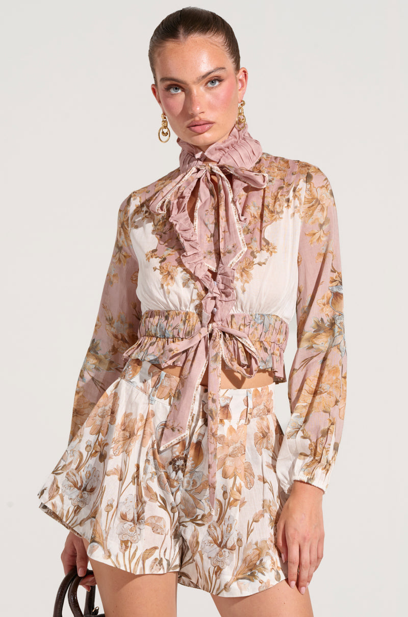 A person stands with a serious expression, wearing the stylish Lucid Dreams Floral Tie Neck Blouse, distinguished by its ruffled high neck and long sleeves, paired with matching high-waisted shorts. They accessorize with gold earrings and hold a small dark handbag against a neutral background.
