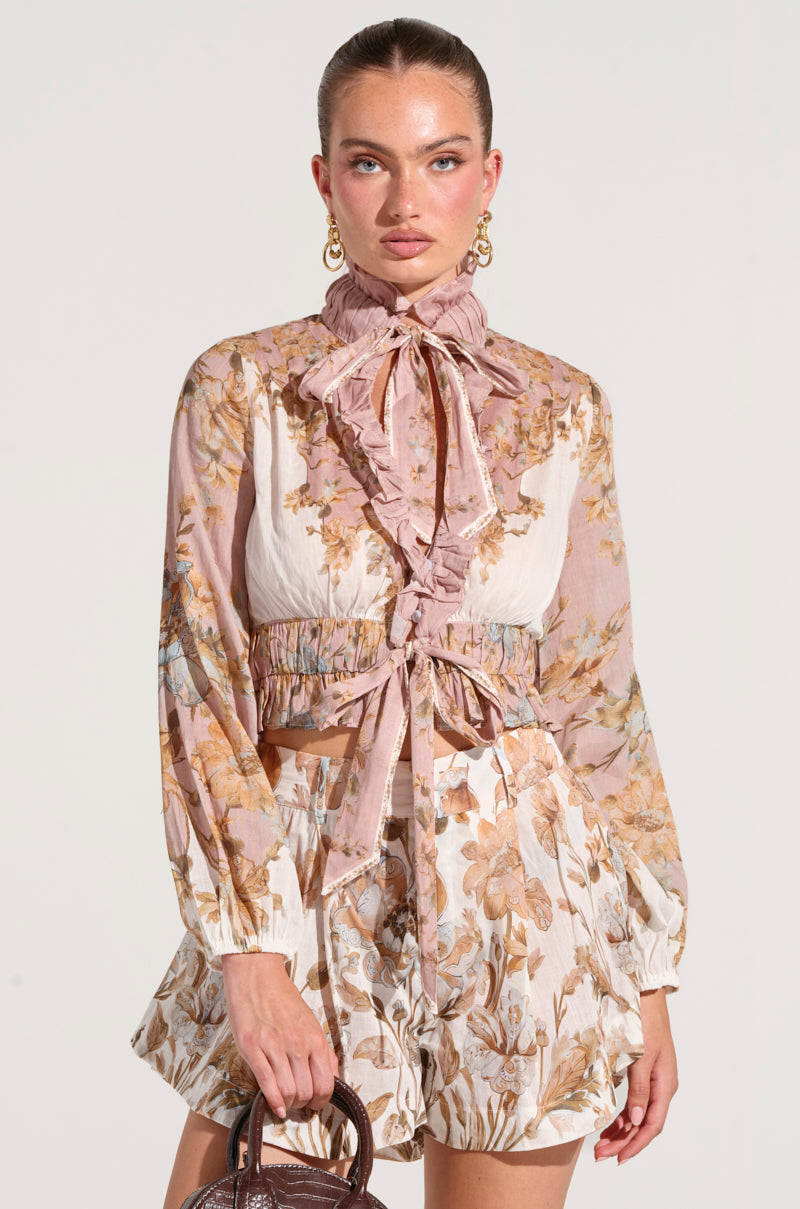 A woman with her hair pulled back wears a Lucid Dreams Floral Tie Neck Blouse with long sleeves and a high ruffled neck, paired with matching high-waisted shorts. She carries a small brown bag and has gold hoop earrings. The background is plain white.