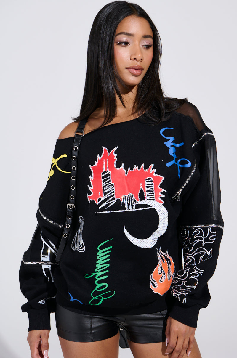 A woman with long black hair models the DEEP THOUGHTS OVERSIZED GRAPHIC SWEATSHIRT featuring colorful abstract designs, including a cityscape and flames. She pairs it with black leather shorts and a black strap, posing gracefully against a light gray background.