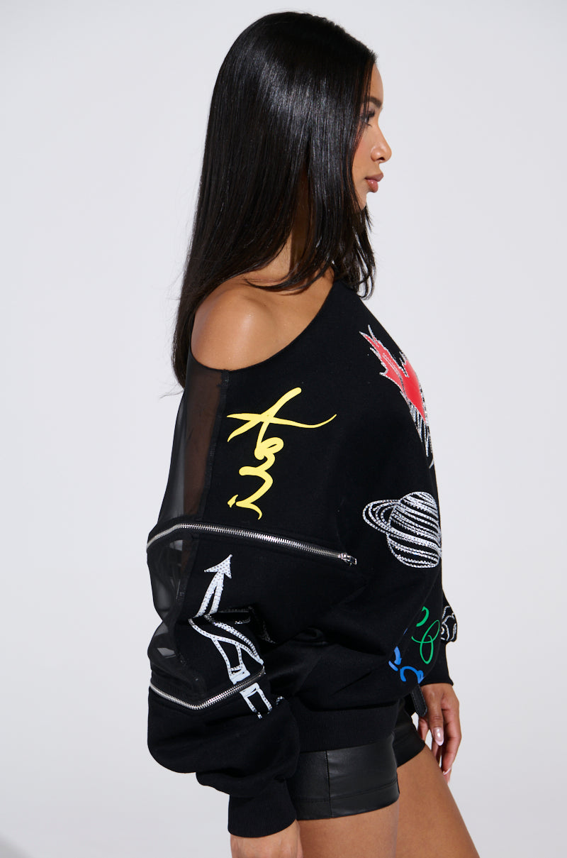 A woman with long dark hair is in profile, highlighting the DEEP THOUGHTS OVERSIZED GRAPHIC SWEATSHIRT featuring colorful designs like a planet and arrows. The cotton-blend fabric enhances the vibrant look. She's wearing black shorts against a plain white background.