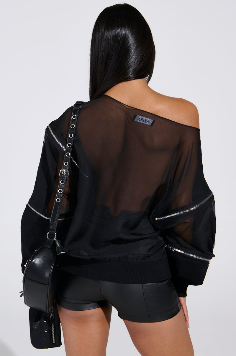 With long dark hair, a person is seen from behind wearing the DEEP THOUGHTS OVERSIZED GRAPHIC SWEATSHIRT, featuring zipper details and rhinestone prints, paired with black shorts. They carry a black leather bag over one shoulder. The sweatshirt casually drapes off one shoulder for a stylish look.