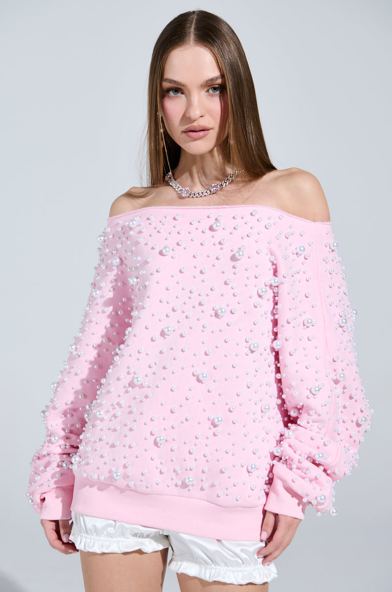With long brown hair, a person wears the PEARL MADNESS OFF THE SHOULDER SWEATSHIRT in pink, cozy cotton-blend with pearl beads, paired with white ruffled shorts and a silver necklace. They pose against a plain background.