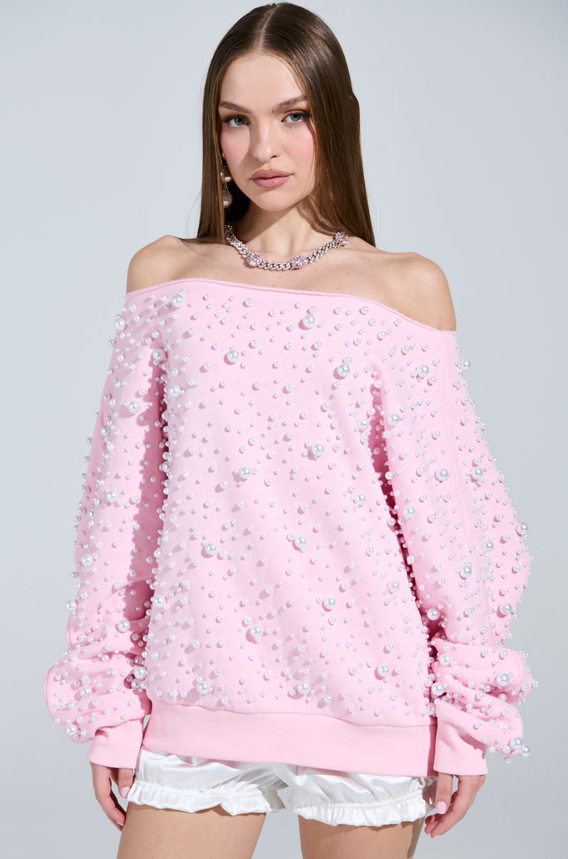 A person with long hair wears the PEARL MADNESS OFF THE SHOULDER SWEATSHIRT, a cozy cotton-blend pink top adorned with pearl beads, and white shorts. They have a neutral expression and accessorize with a chain necklace. The background is plain and light-colored.