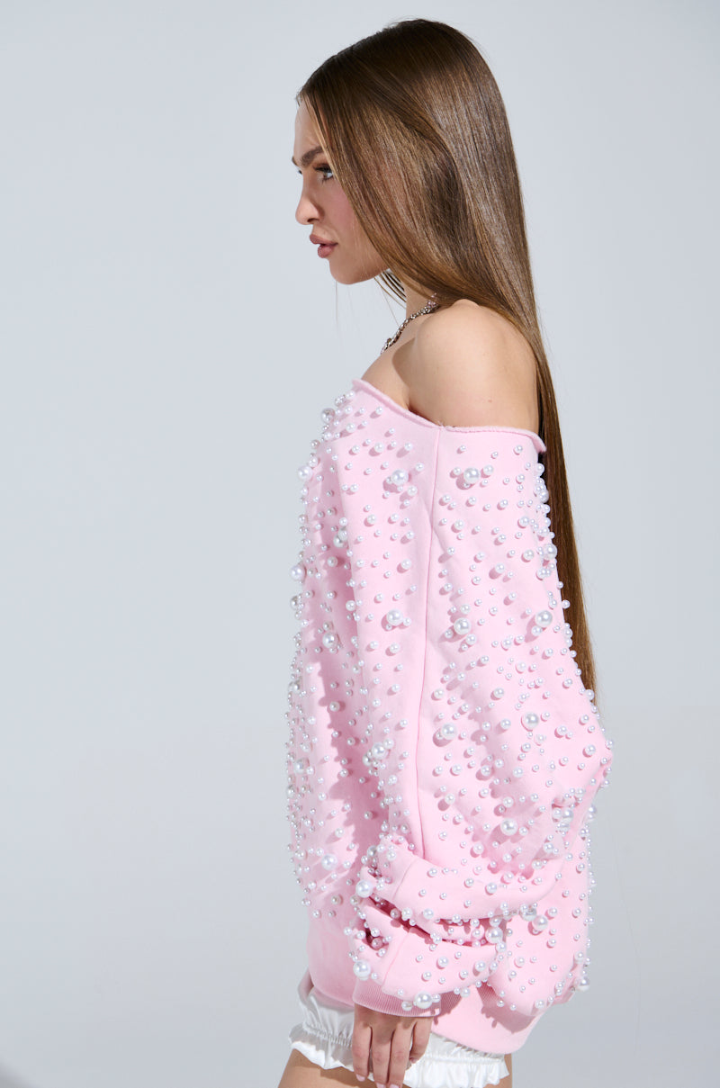 With long straight hair, a person models sideways against a plain gray background, wearing the PEARL MADNESS OFF THE SHOULDER SWEATSHIRT with cozy cotton-blend material and pearl-like embellishments.