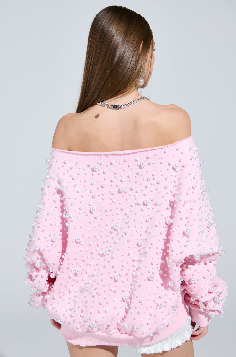 A woman with long brown hair models the cozy PEARL MADNESS OFF THE SHOULDER SWEATSHIRT adorned with pearl beads. Facing away, she elegantly highlights her silver necklace and earrings against a light gray backdrop.
