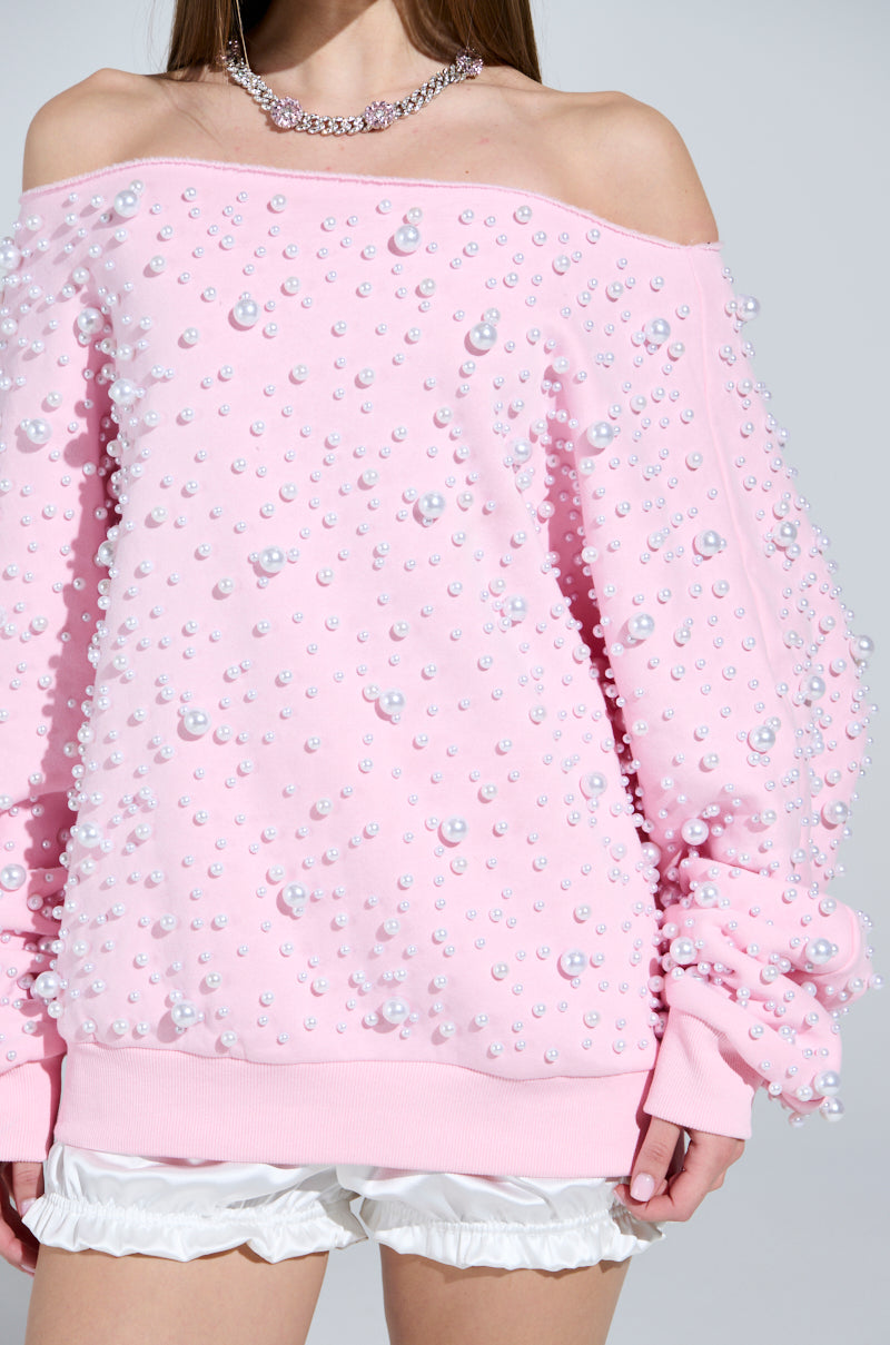 A person dons the PEARL MADNESS OFF THE SHOULDER SWEATSHIRT, a cozy cotton-blend piece adorned with pearl beads, paired with white ruffled shorts. The focus is on the detailed upper body attire and a delicate necklace.