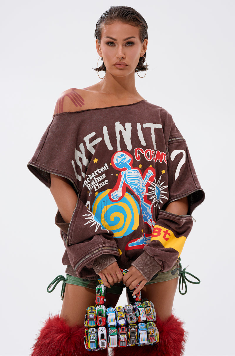 A woman with slicked-back hair wears the INFINITE COSMOS OVERSIZED GRAPHIC SWEATSHIRT, paired with green shorts, red furry boots, and a toy car-embellished handbag, standing against a plain white background.