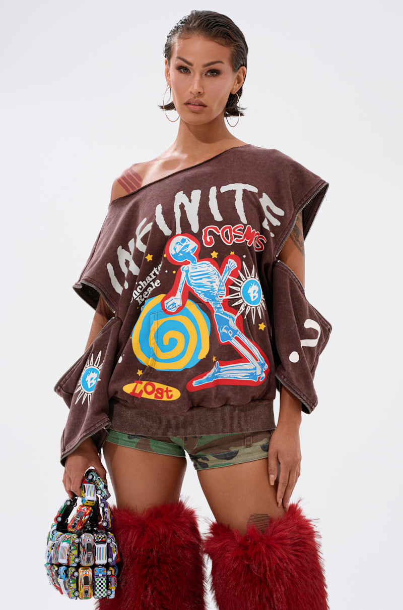 A woman poses in the INFINITE COSMOS OVERSIZED GRAPHIC SWEATSHIRT, styled with camo shorts, bright red furry boots, and a multicolored handbag against a plain light background.