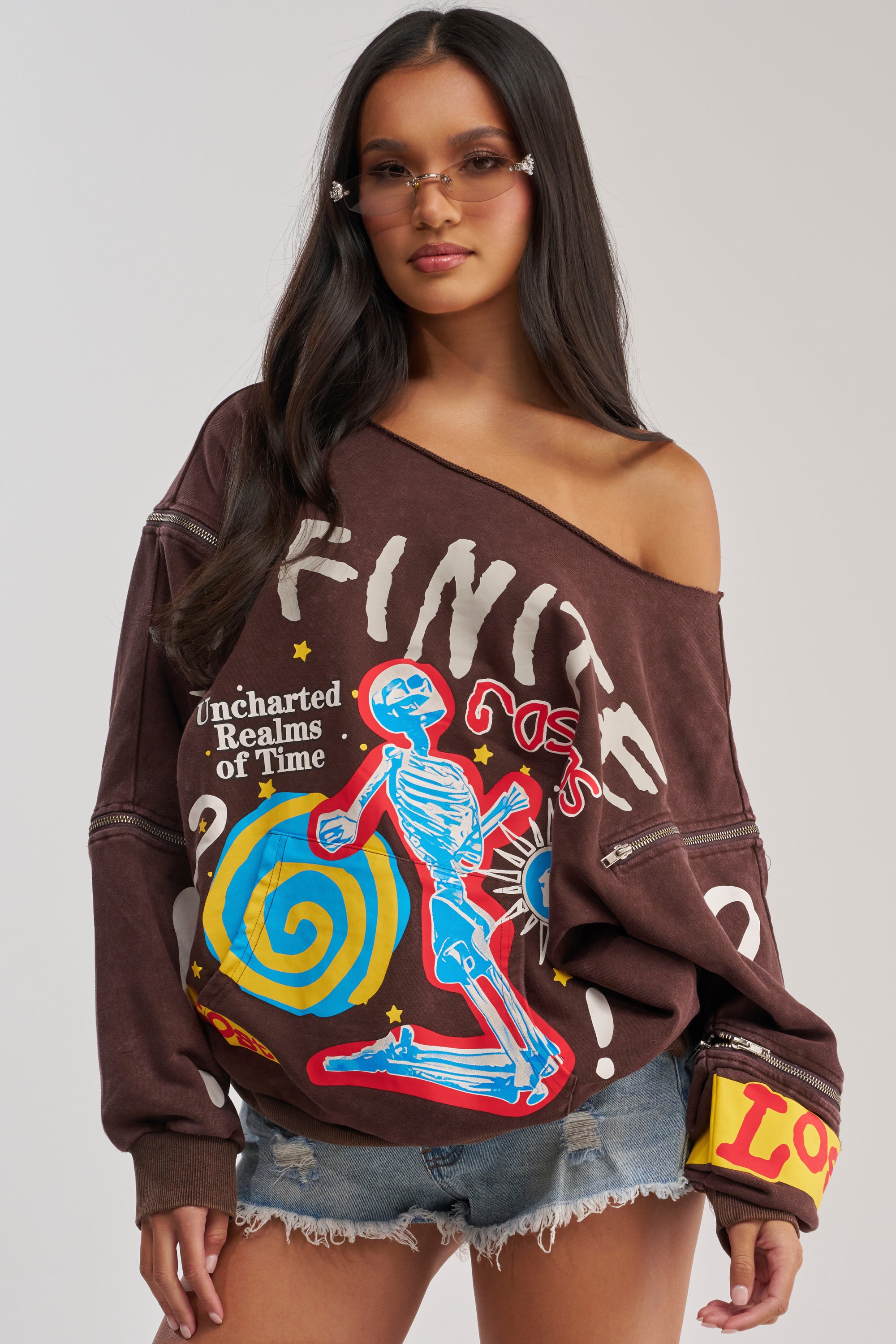 A woman with long, dark hair wears angular sunglasses and the INFINITE COSMOS OVERSIZED GRAPHIC SWEATSHIRT—an off-the-shoulder brown top with colorful graphics and text—paired with distressed denim shorts.
