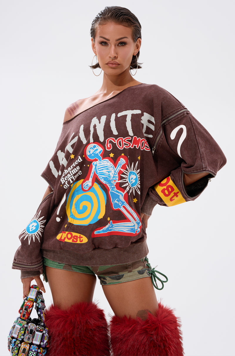 A woman poses confidently in the INFINITE COSMOS OVERSIZED GRAPHIC SWEATSHIRT with colorful artwork and text, camo shorts, large hoop earrings, red furry boots, and a small patterned bag against a plain white background.