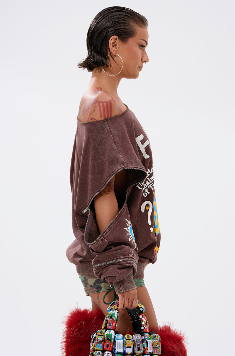 A person with slicked-back hair, large hoop earrings, and visible tattoos wears an INFINITE COSMOS OVERSIZED GRAPHIC SWEATSHIRT, camo shorts, and holds a colorful handbag featuring red fur and toy car handles.