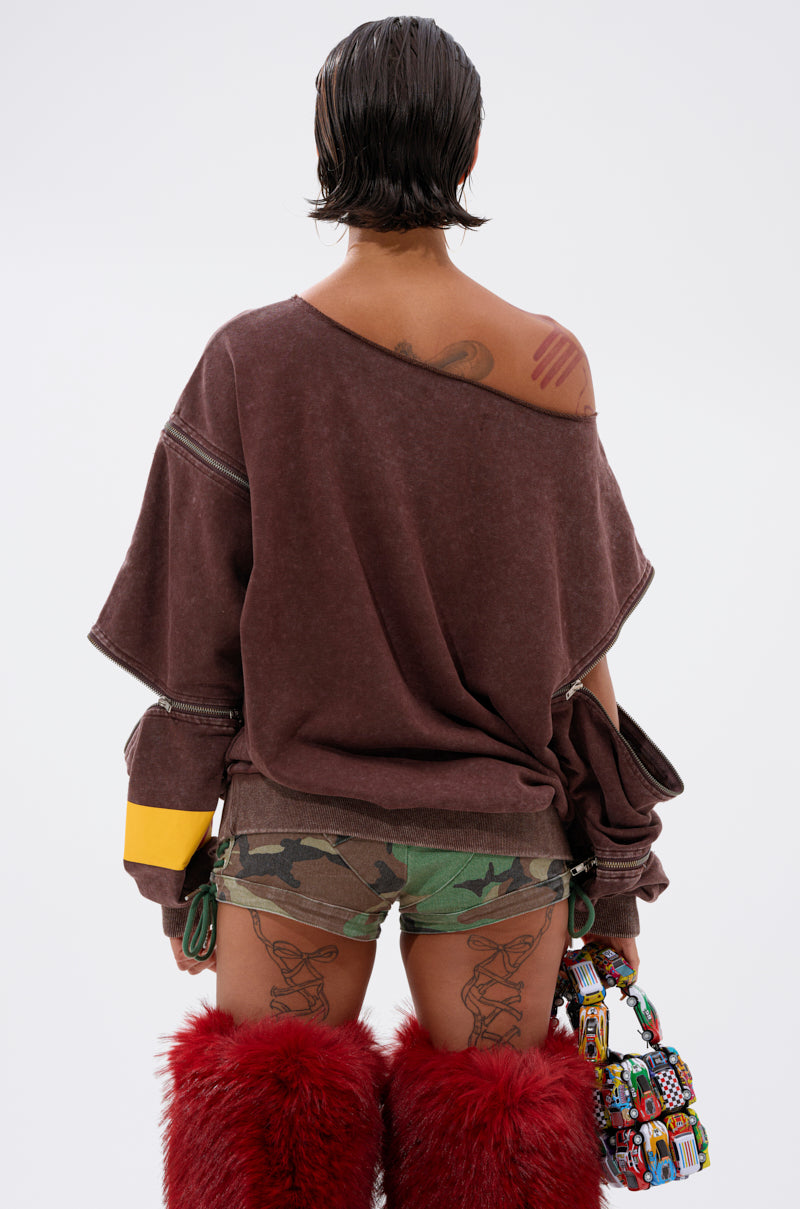 A person with short dark hair faces away, wearing an INFINITE COSMOS OVERSIZED GRAPHIC SWEATSHIRT, green camo shorts, red furry boots, and holding a colorful beaded bag. Tattoos are visible on their arms and legs.