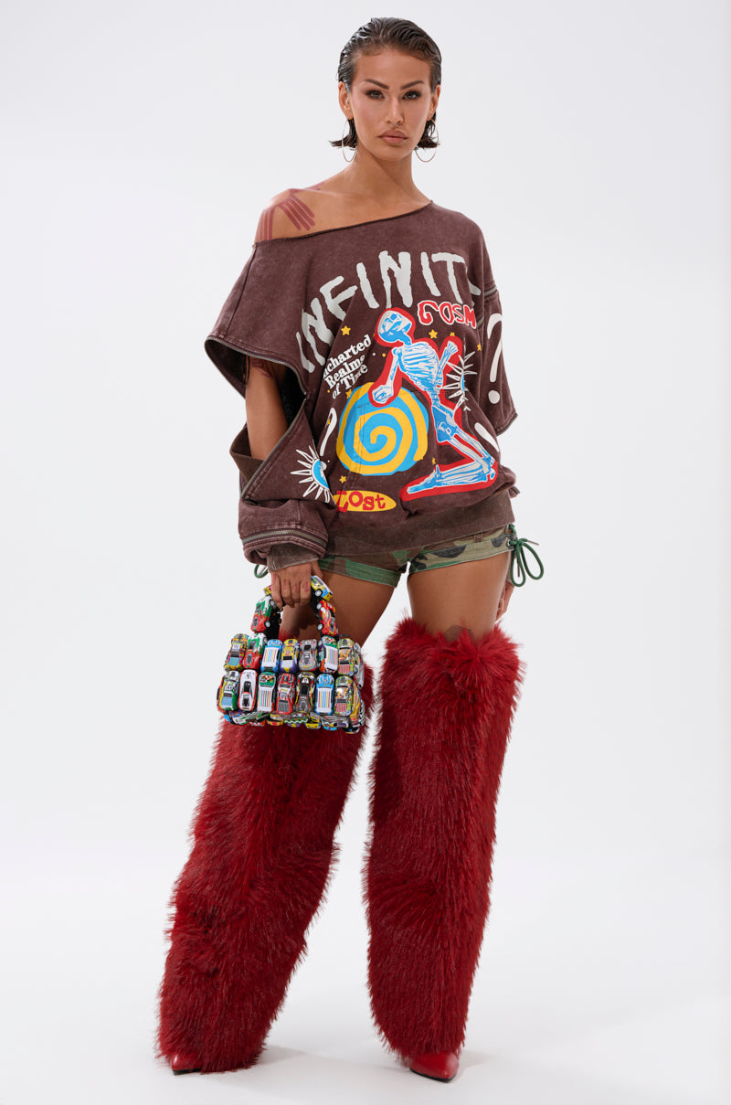 A person in an INFINITE COSMOS OVERSIZED GRAPHIC SWEATSHIRT, green shorts, bright red furry thigh-high boots, and holding a colorful beaded handbag stands against a plain white background.