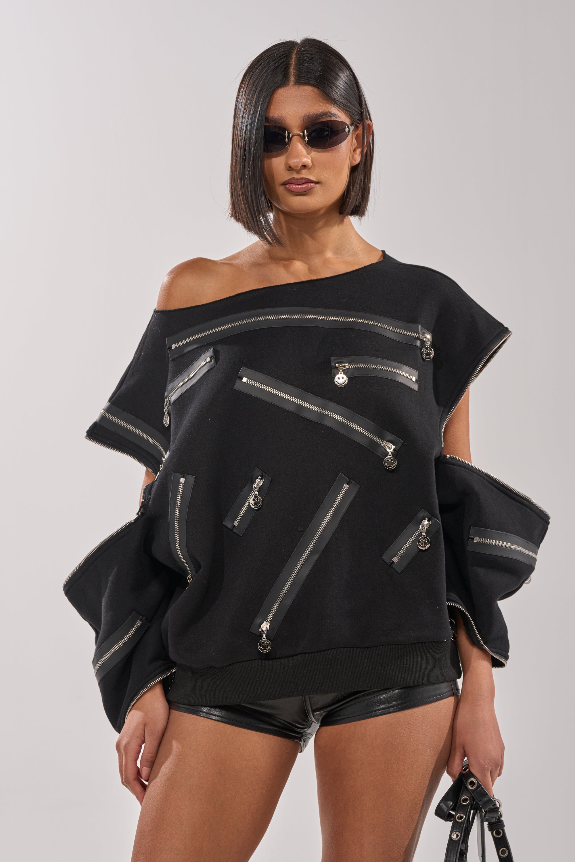 A woman with a sleek bob and sunglasses stuns in the UN-ZIP ME OFF THE SHOULDER SWEATSHIRT, styled with black leather shorts and a black bag featuring metal rings for an extra edgy vibe. 0