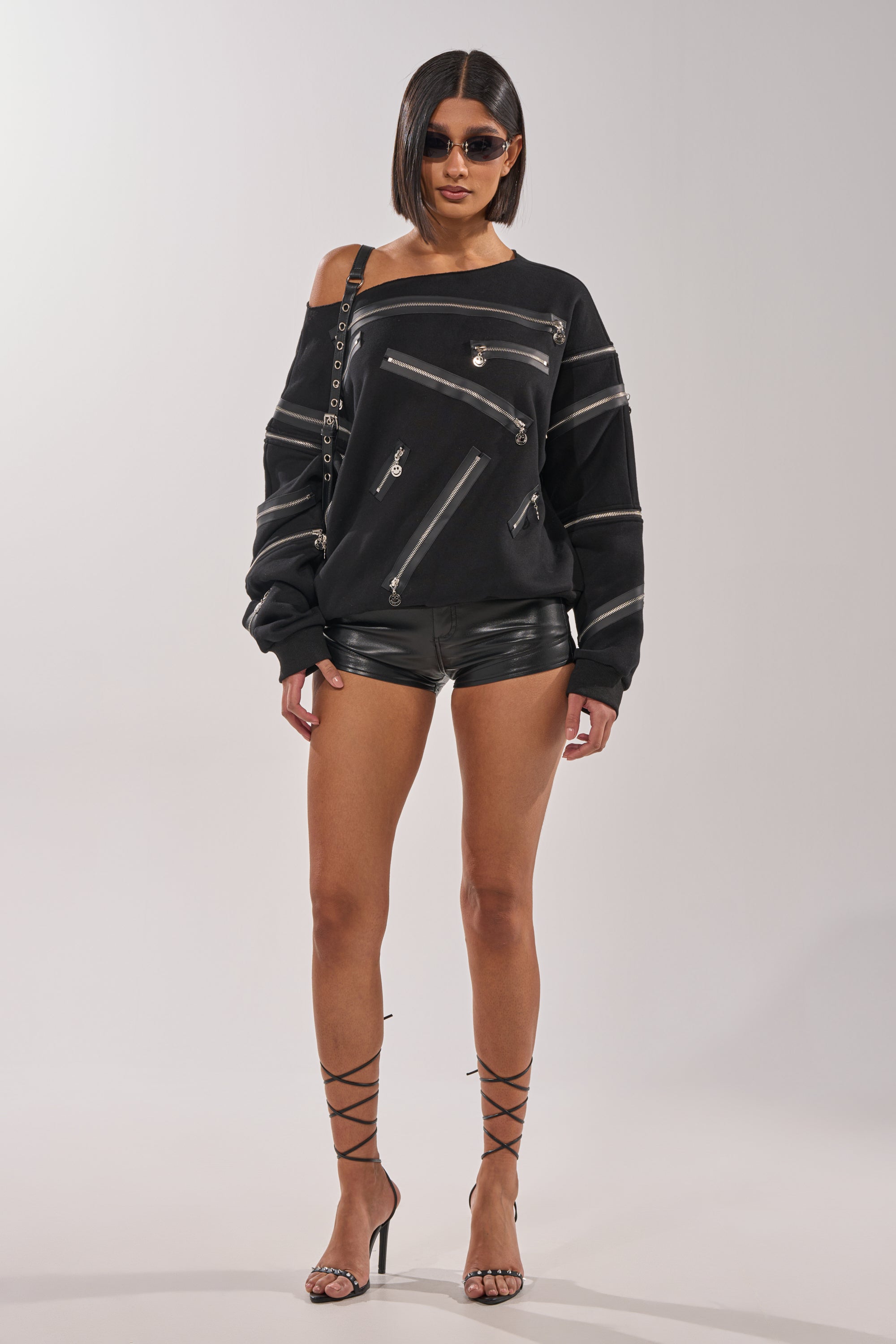 A woman wears the UN-ZIP ME OFF THE SHOULDER SWEATSHIRT in black, paired with black leather shorts, strappy high heels, sunglasses, and a black shoulder bag, standing against a plain light background. 9