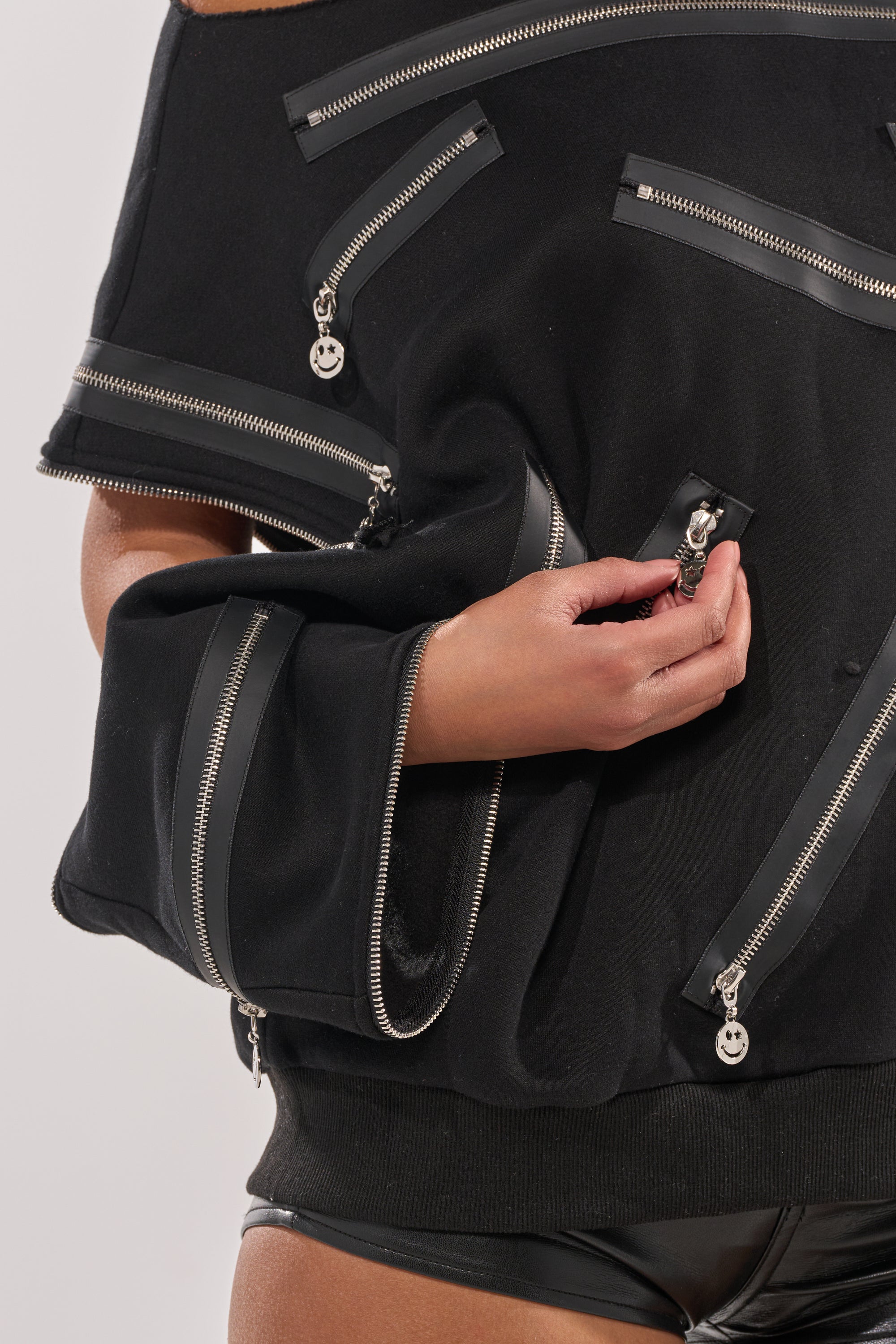 Wearing the UN-ZIP ME OFF THE SHOULDER SWEATSHIRT in black with several silver zippers, some partly open and holding one zipper, a person pairs it with black shorts and stands against a plain light background. 12