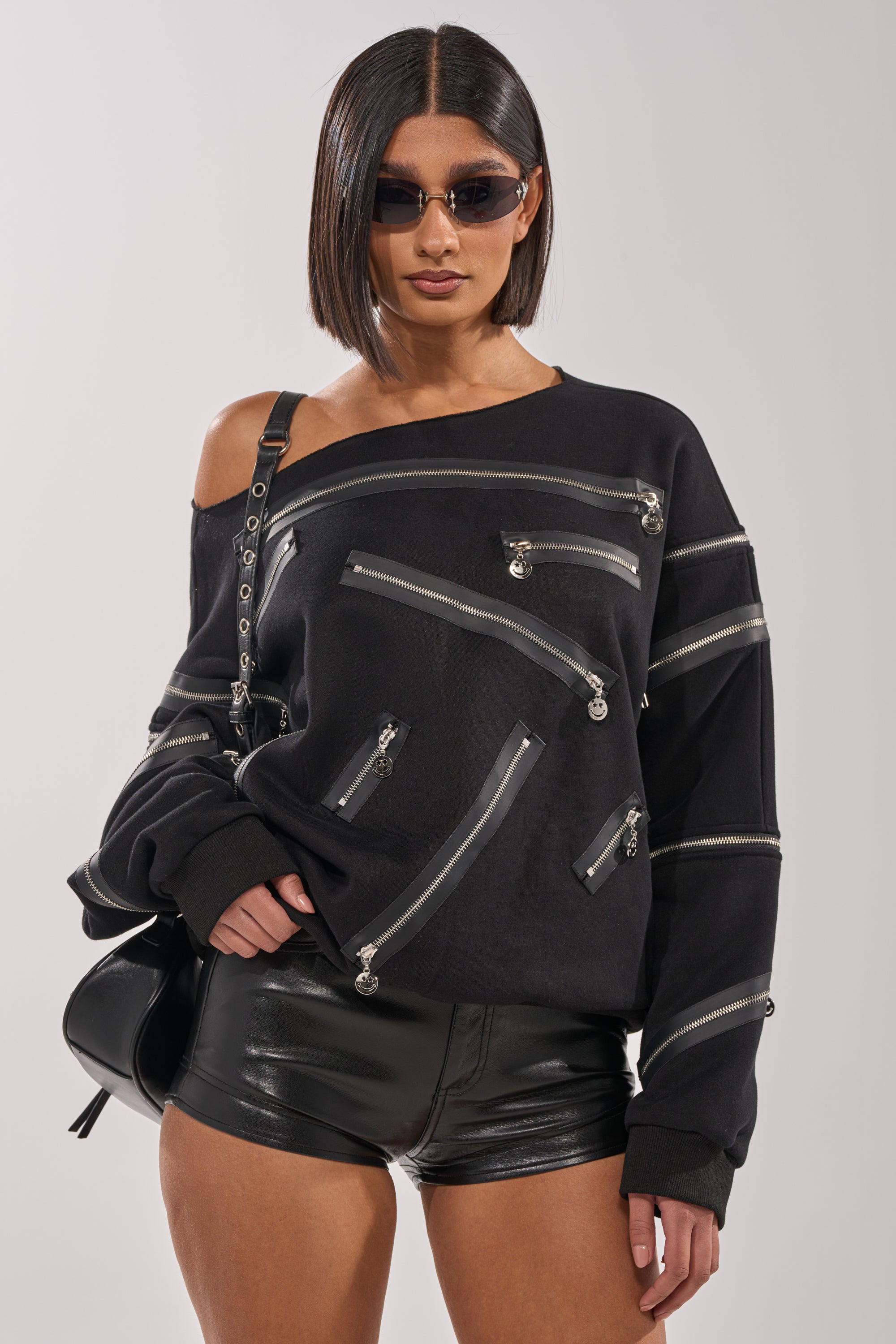 A woman with a sleek bob and sunglasses wears the UN-ZIP ME OFF THE SHOULDER SWEATSHIRT, black leather shorts, and carries a black shoulder bag—capturing effortless streetwear style. 1